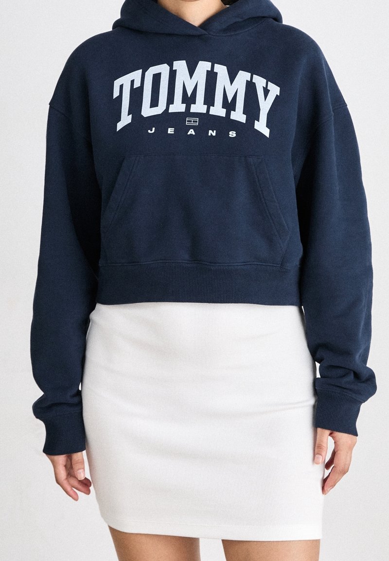Felpa corta blu navy con la scritta "TOMMY JEANS" in bianco, dotata di tasca frontale e polsini a costine, abbinata a una gonna bianca a costine.