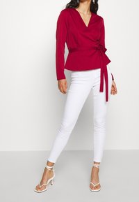 Top rojo envuelto con mangas largas, escote en V, cintura ceñida con un lazo, combinado con jeans skinny blancos y tacones blancos con tiras.