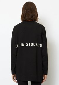 Sudadera negra hecha de material suave, con un corte relajado. Incluye el texto blanco "EST. EN ESTOCOLMO" en la parte trasera. Mangas largas, puños acanalados.