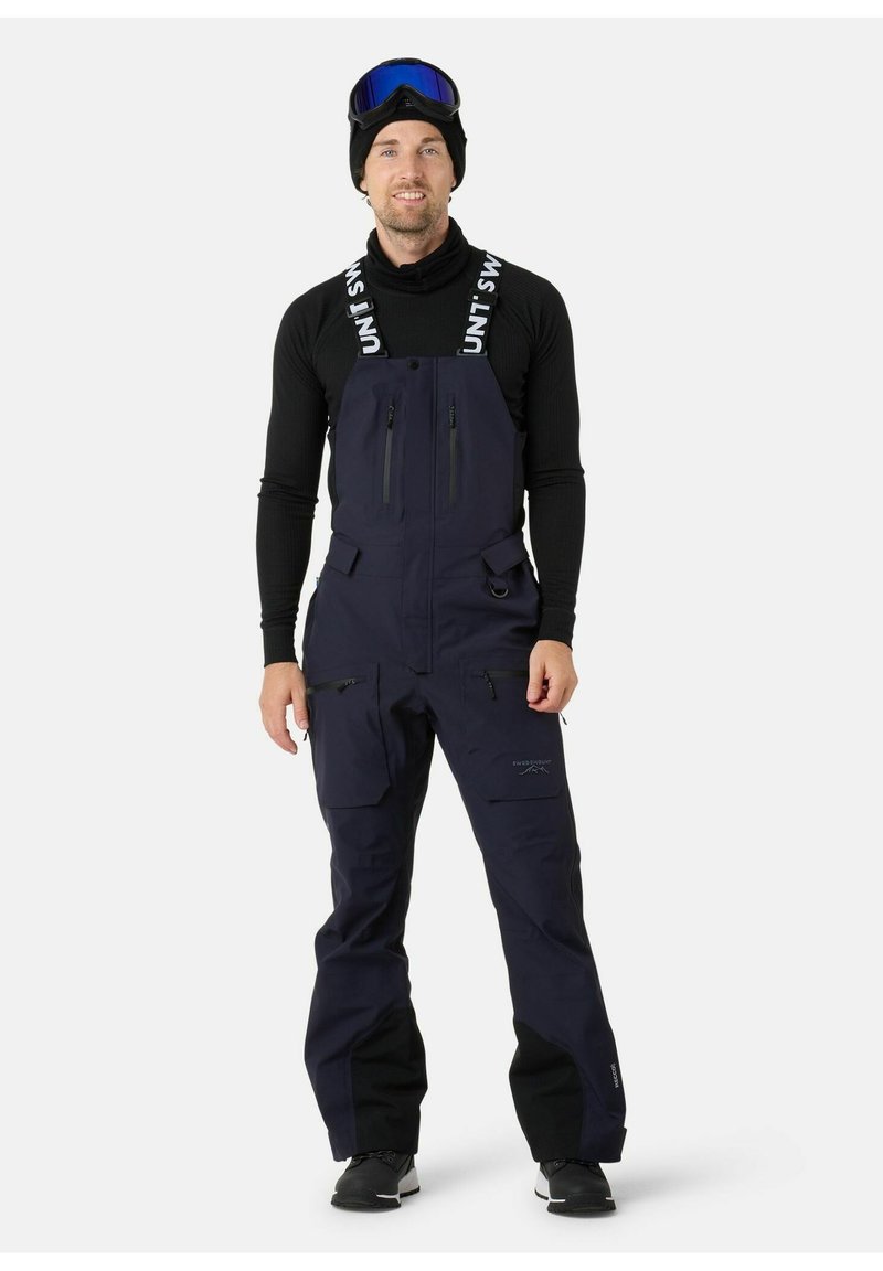 Tuta da sci blu navy con spalline regolabili, tasche con zip e un design resistente alle intemperie. Indossata con una maglietta nera a maniche lunghe e occhiali da sci.