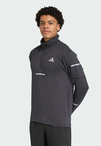 Pull à col zippé mi-haut gris foncé, à manches longues, avec des accents réfléchissants, une texture côtelée et un logo brodé sur la poitrine et la poche de la manche.