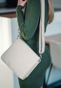 Béžová kožená crossbody taška s nastavitelným popruhem, obdélníkový tvar, embosované logo a hladká textura. Nosená k zeleným žebrovaným šatům.