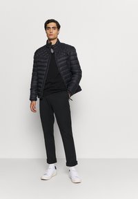 Gequiltede zwarte pufferjack met een hoge kraag, gecombineerd met een zwart shirt en opgerolde zwarte broek. Witte sneakers maken de look compleet.