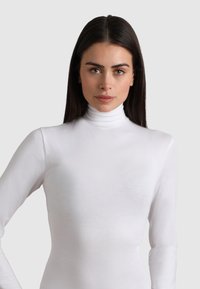 Donna con lunghi capelli scuri che indossa un dolcevita bianco, in piedi contro uno sfondo grigio chiaro uniforme, che guarda direttamente di fronte a sé.