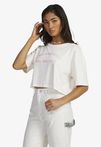 White cropped t-shirt with pink text "SUNSETS AND MISERY CALL 1800 - RVC," paired with white pants featuring a black and white striped accent.