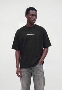 Young man wearing a black oversized T-shirt with "DROPSIZE" text and faded gray jeans, standing with one hand in his pocket.
