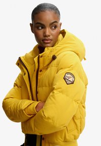 Gul pufferjakke med hætte, der har en brun lap på ærmet, trykknapper og en glat tekstur. Lynlås og syninger er synlige.