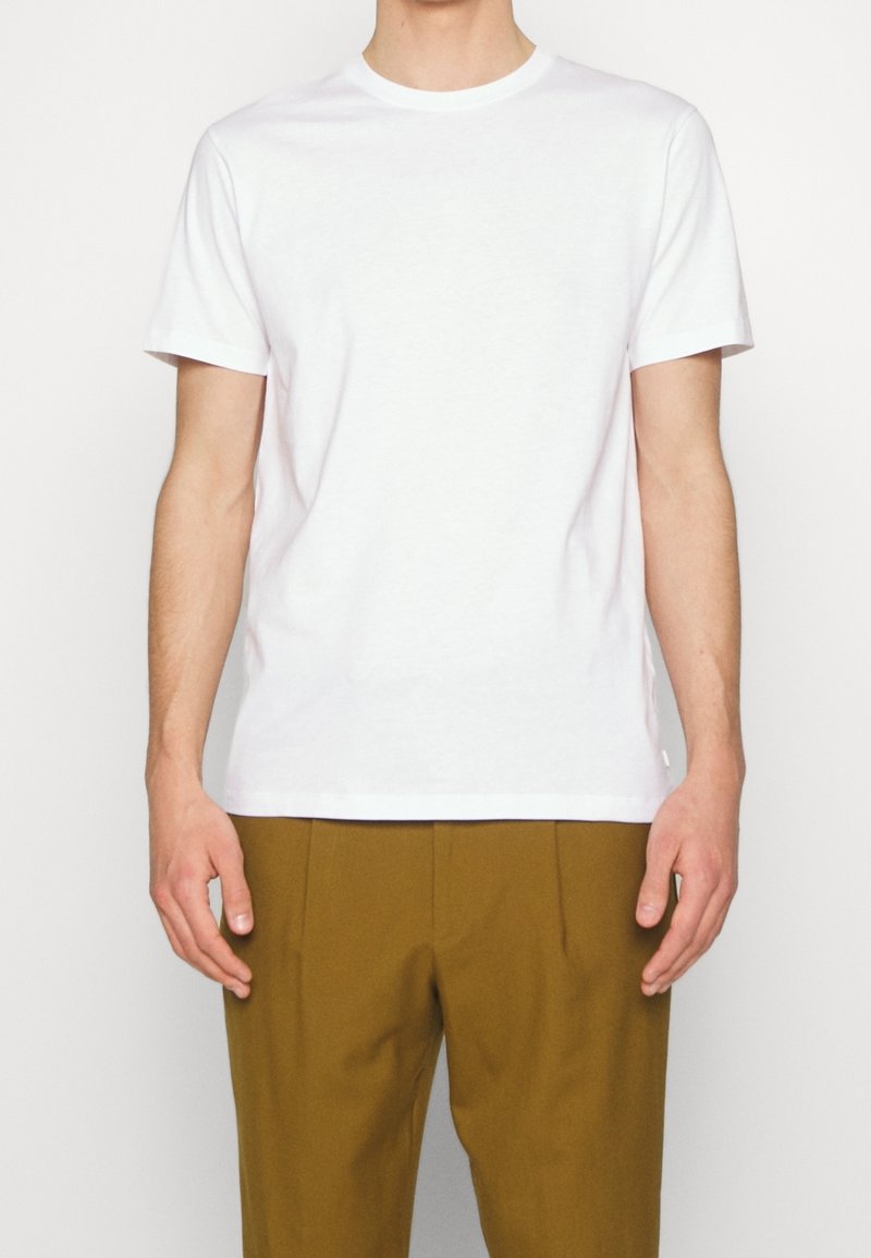 T-shirt en coton blanc à manches courtes et encolure ras du cou, porté par une personne. Assorti avec un pantalon plissé marron.