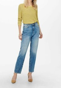 Maglione giallo a maglia, jeans a vita alta di colore blu chiaro, gamba dritta, finitura sbiadita, look usurato, abbinato a tacchi beige su uno sfondo bianco.