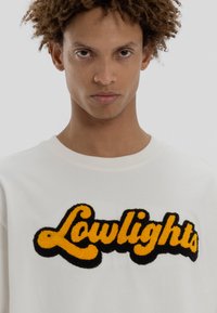 Νέος άνθρωπος με σγουρά μαλλιά που φοράει λευκό πουλόβερ με κίτρινα και μαύρα κεντημένα γράμματα "Lowlights" μπροστά.