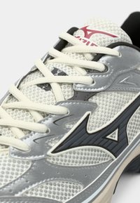 White and gray athletic shoe with mesh texture, cream laces, and black and red logo details on the tongue and side.