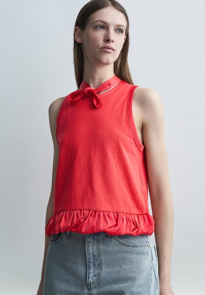 Femme portant un haut rouge sans manches avec un nœud au niveau de l'encolure et un ourlet froncé, associée à un jean en denim bleu clair, sur un fond uni.