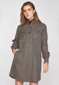 Vestito camicia marrone a quadri con maniche lunghe, chiusura frontale con bottoni e due tasche sul petto. Caratterizzato da una silhouette rilassata e tasche laterali.