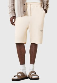 Pantaloni corti di cotone beige con vita elastica e tasche laterali, con un discreto testo del logo, abbinati a sandali marroni e calzini bianchi.