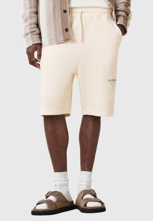 Beige katoenen shorts met een elastische tailleband en zijzakken, voorzien van subtiele logotext, gecombineerd met bruine sandalen en witte sokken.