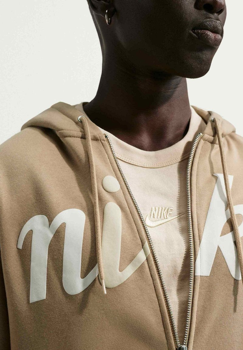 Person wearing a beige zip-up Nike hoodie over a matching T-shirt with Nike logo, shown from shoulders to mouth.