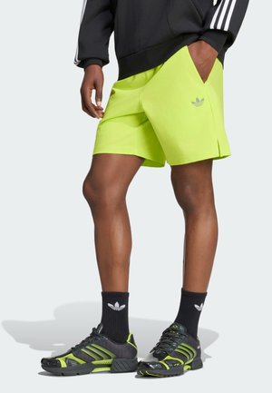 Osoba nosząca neonowo-zielone szorty, czarne buty Adidas z zielonymi akcentami, czarne skarpety oraz czarną bluzkę z długim rękawem w białe paski.