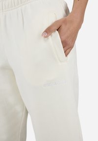 Lichtbeige trainingsbroek met een relaxte pasvorm, voorzien van een zijzak en een subtiele witte geborduurde logo nabij de heup. Zacht, gestructureerd materiaal.