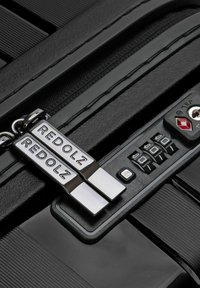 Gros plan sur des tirettes de fermeture éclair de valise noire étiquetées "REDOLZ" sécurisées dans un cadenas à combinaison à trois chiffres avec un trou de serrure TSA.