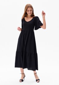 Schwarzes Maxikleid mit kurzen, ausgestellten Ärmeln und einem gestuften Rüschensaum. Glattes Gewebe mit tailliertem Oberteil und V-Ausschnitt.