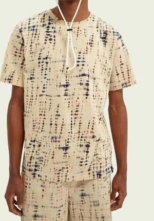 Uomo con camicia beige a maniche corte e pantaloncini abbinati con motivo astratto a pois blu, marrone e nero.