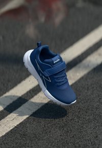 Chaussure de sport bleue avec un dessus en mesh, semelle blanche, bande réglable et lacets ; présente des accents blancs diagonaux sur le côté.