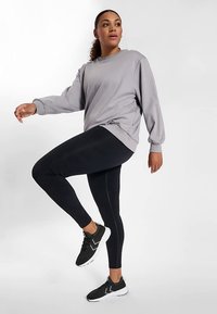 Grijs shirt met lange mouwen, zwarte legging en zwarte sportschoenen. Het shirt heeft een losse pasvorm; de legging is nauwsluitend met een geribbelde textuur.