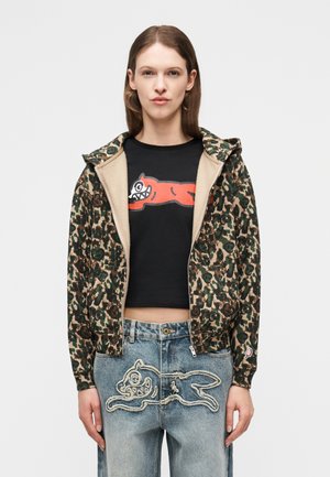 Γυναίκα που φοράει φούτερ με φερμουάρ και μοτίβο παραλλαγής, μαύρο κοντό τοπ με κόκκινο καρτούν ζώο και τζιν με ραμμένο σχέδιο καρτούν ζώου.