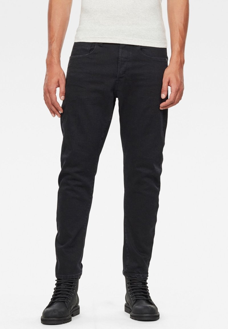 Vaqueros slim-fit negros con una textura suave, cinco bolsillos y pasadores para cinturón, combinados con botas negras de cordones. Diseño sencillo, sin patrones.