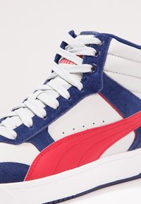 Vysoké tenisky s bílou koženou základnou, modrými semišovými akcenty a červeným syntetickým logem na boku. Šňůrky jsou světle šedé. Ploché gumové podrážky.