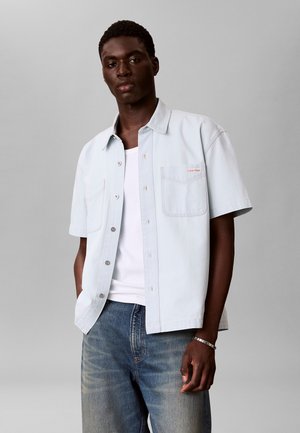 Junger Mann trägt ein hellblaues kurzärmeliges Hemd über einem weißen T-Shirt und blauen Jeans, steht vor einem schlichten grauen Hintergrund.