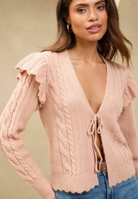 Šviesiai rožinė megzta kardiganė su banguotomis rankovėmis, pynėmis, giliu V formos iškirpimu ir kaklo raiščiu priekyje. Puošnus apatinis kraštas.