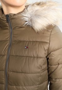 Chaqueta acolchada verde oliva con cremallera, que cuenta con una capucha de piel sintética y un pequeño acento de logo. El material parece suave con detalles de costura horizontal.