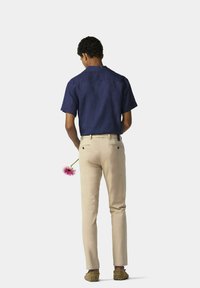 Marineblaues Kurzarmhemd, beige angepasste Hose und grüne Schuhe. Eine pinkfarbene Blume wird hinter dem Rücken gehalten. Einfarbiger Hintergrund.
