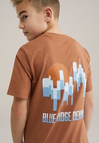 Ruda medvilninė marškinėliai su mėlynu miesto panorama grafiku ir dideliu oranžinės saulės piešiniu ant nugaros. Tekstas skaito "BLUE RIDGE DENIM."