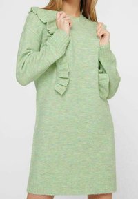Zielona dzianinowa sukienka o miękkiej fakturze, z falbanami na ramionach i luźnym kroju, sięgająca nieco powyżej połowy uda.