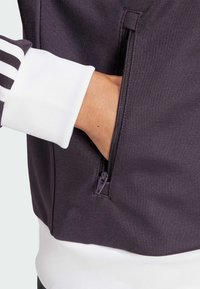 Bolsillo con cremallera de una chaqueta de color púrpura oscuro con tejido texturizado, que presenta puños de canalé blancos y una cinturilla blanca. Mano visible dentro del bolsillo.