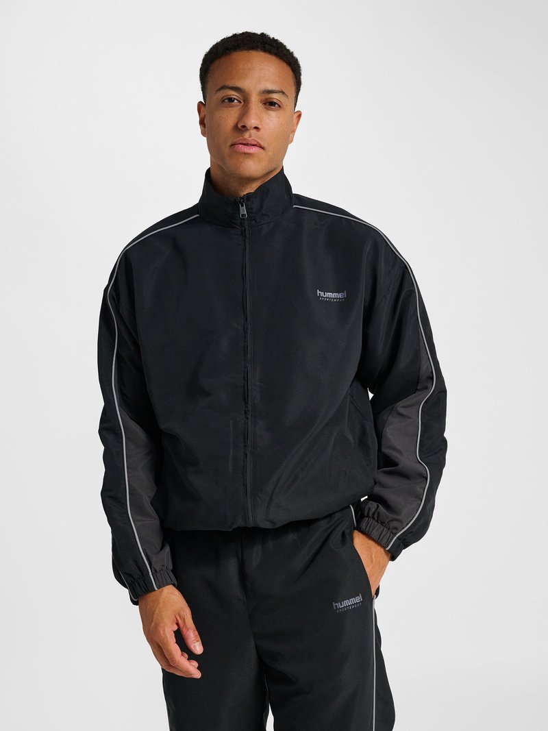 Mann trägt schwarze Sportjacke mit Reißverschluss und passende Hose mit dezenter reflektierender Paspel vor einfarbigem Hintergrund.
