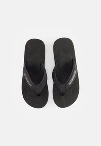 Des tongs noires avec des semelles en caoutchouc et des sangles texturées. Elles présentent un logo embossé et un design minimaliste. Plate et de style entre-doigts.