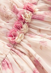 Robe fleurie en tissu léger rose pastel. Présente des fleurs en tissu roses et crème le long du corsage, avec une texture froncée.