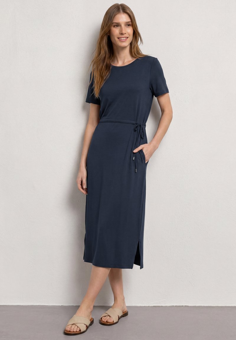 Femme debout dans une robe midi bleu marine à manches courtes, fente latérale, ceinture à la taille et sandales beiges à enfiler contre un mur blanc uni.