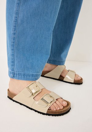 Beige lackskor med två justerbara spännen, korkfotsäng och svart texturerad sula, tillsammans med ljusblå denimbyxor.