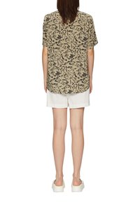 Kurzärmliges, hellbeiges Hemd mit einem schwarzen Blumenmuster, lockere Passform und runder Saum, kombiniert mit weißen Leinen-Shorts und flachen Schuhen.