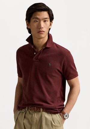 Burgundská polo košile s límečkem a dvěma knoflíky, se malým zeleným logem. Doplněná světlými khaki kalhotami a hnědým koženým páskem.