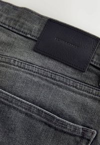 Gros plan sur la poche arrière d'un jean en denim gris avec une étiquette en cuir noir cousue au-dessus, embossée du texte "DENIM SUPPLY".