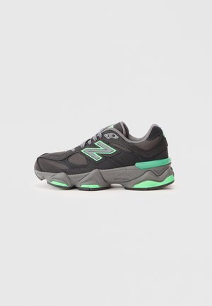 Zwart en grijs atletisch sneaker met groene accenten en "N" logo, meshpanelen en dikke gevoerde zool op een witte achtergrond.