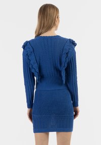 Vestito a maglia blu con spalline a volants, texture a costine e silhouette aderente. Presenta motivi a punto aperto lungo il corpo.