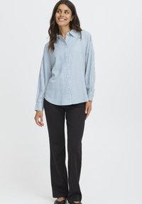 Woman smiling, wearing a light blue and white striped button-up shirt with long sleeves, black straight-leg pants, and black shoes on white background.