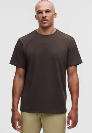 Volwassen man met geschoren hoofd, draagt een effen donkerbruine T-shirt met korte mouwen en licht beige broek tegen een effen achtergrond.