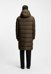 Bruine pufferjas met capuchon, gekenmerkt door een doorgestikt ontwerp en een lange snit. Draagt over zwarte broek, wat een strakke, matte textuur laat zien.